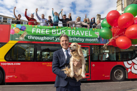 Photo of a man holding a dog standing in front of an open-top bus that has cheering people on the top deck.