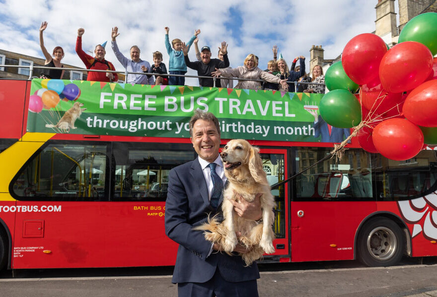 Photo of a man holding a dog standing in front of an open-top bus that has cheering people on the top deck.